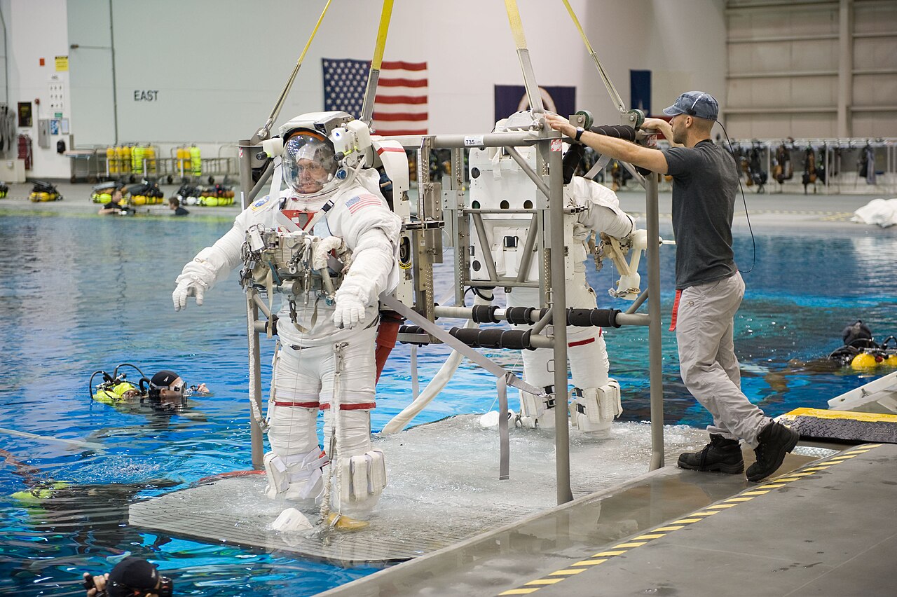 Astronauta de traje espacial sendo preparado na borda da piscina do laboratório, com mergulhadores na água