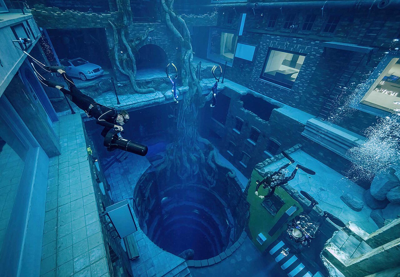 Mergulhadores dentro da Deep Dive Dubai, com cenário de cidade submersa e o poço central que desce até 60 metros